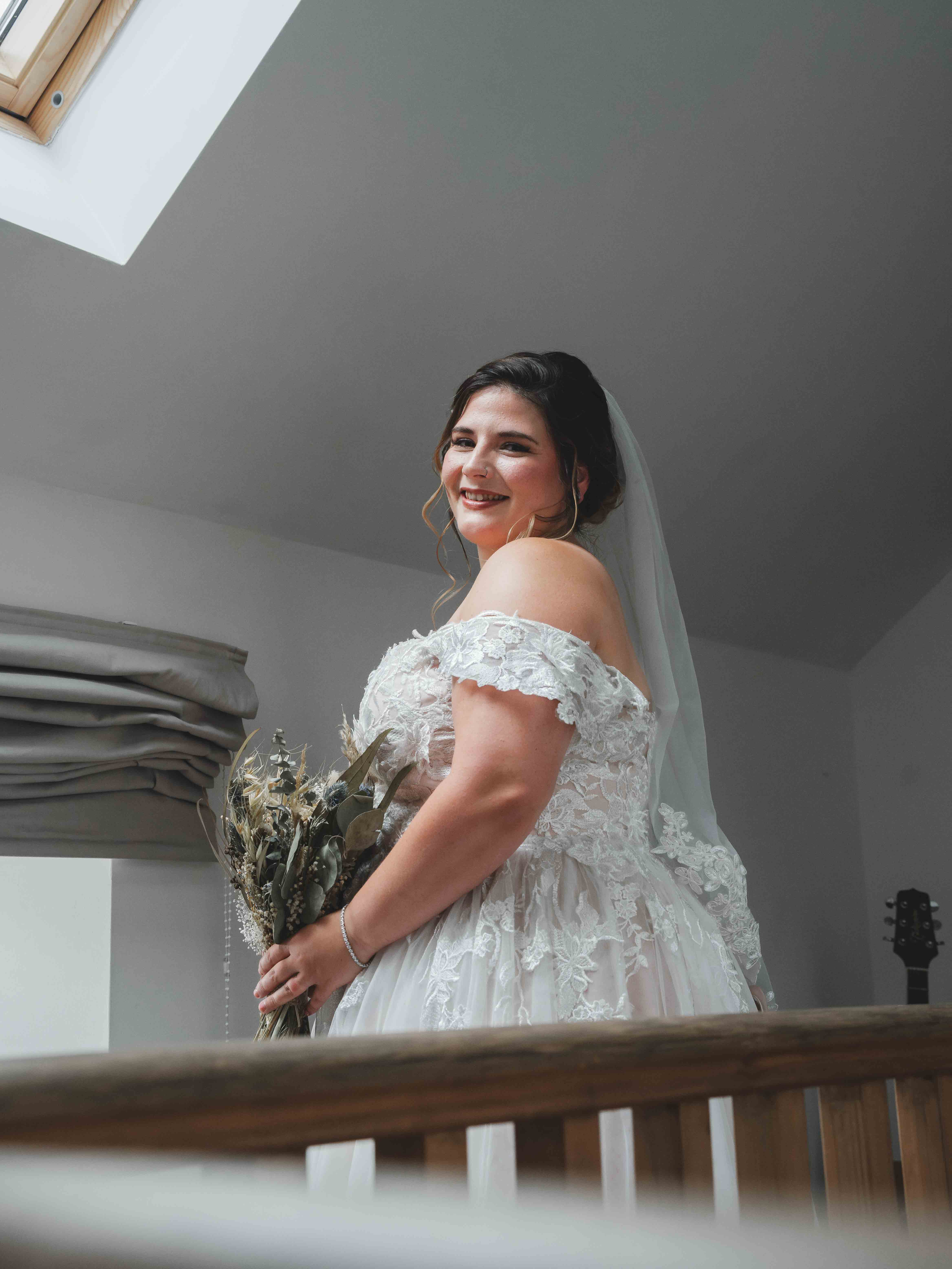 Cessca smiling on the staircase in her off-shoulder lace wedding dress with veil and dried flower bouquet