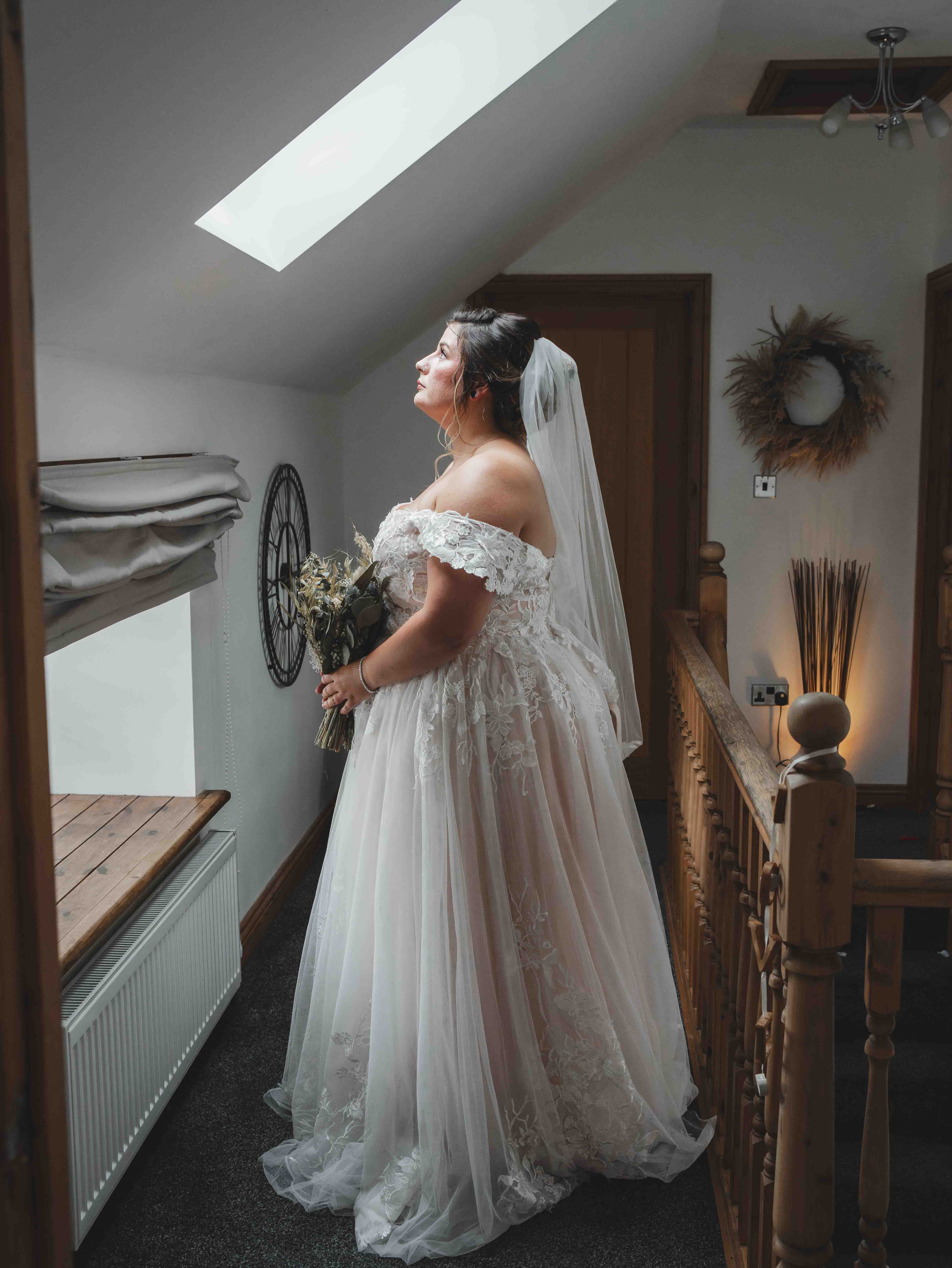 Cessca full-length bridal portrait by a skylight in her lace wedding dress with veil and dried flower bouquet