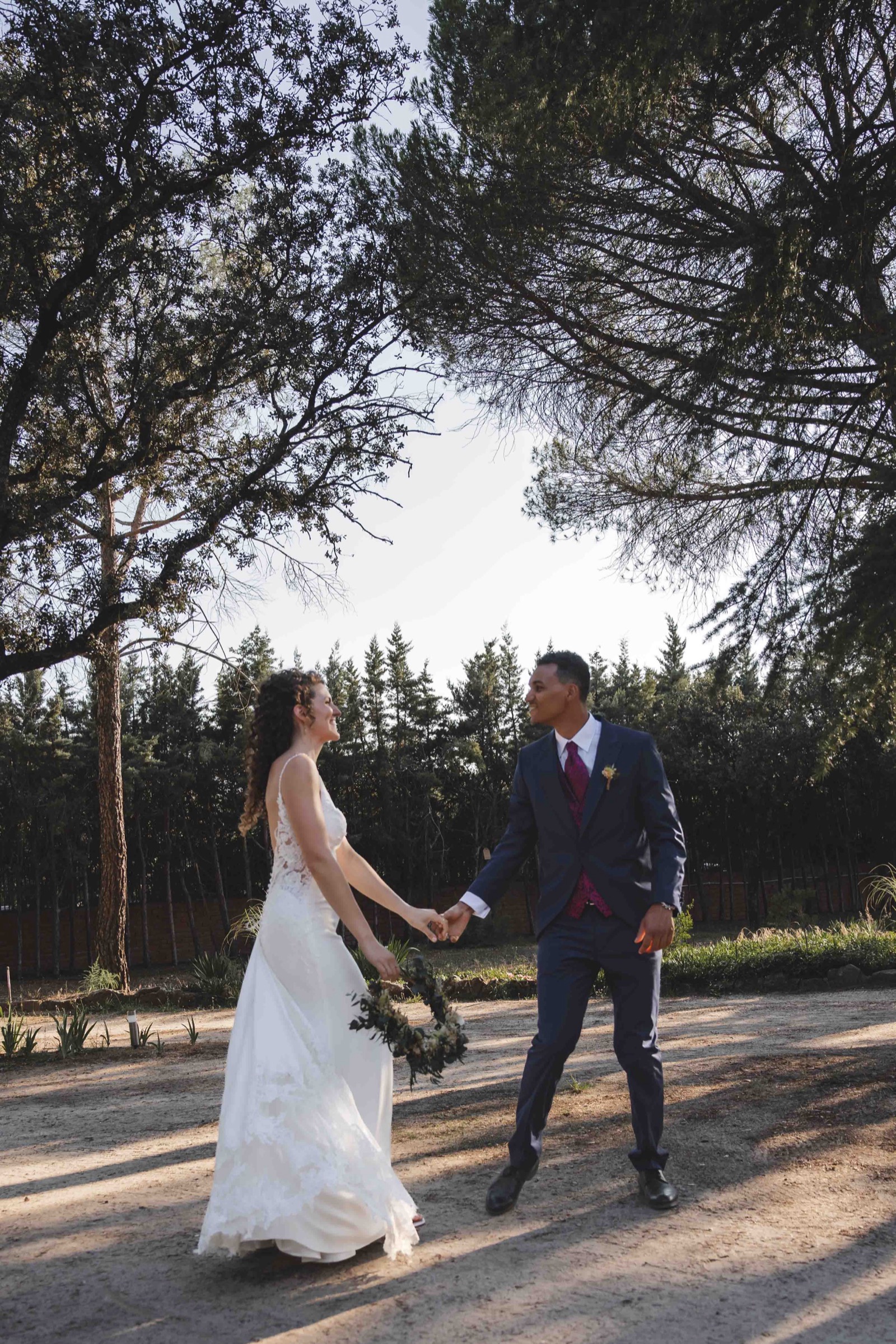 Maria and Leo walking hand in hand along a tree-lined path, golden afternoon light between the pines