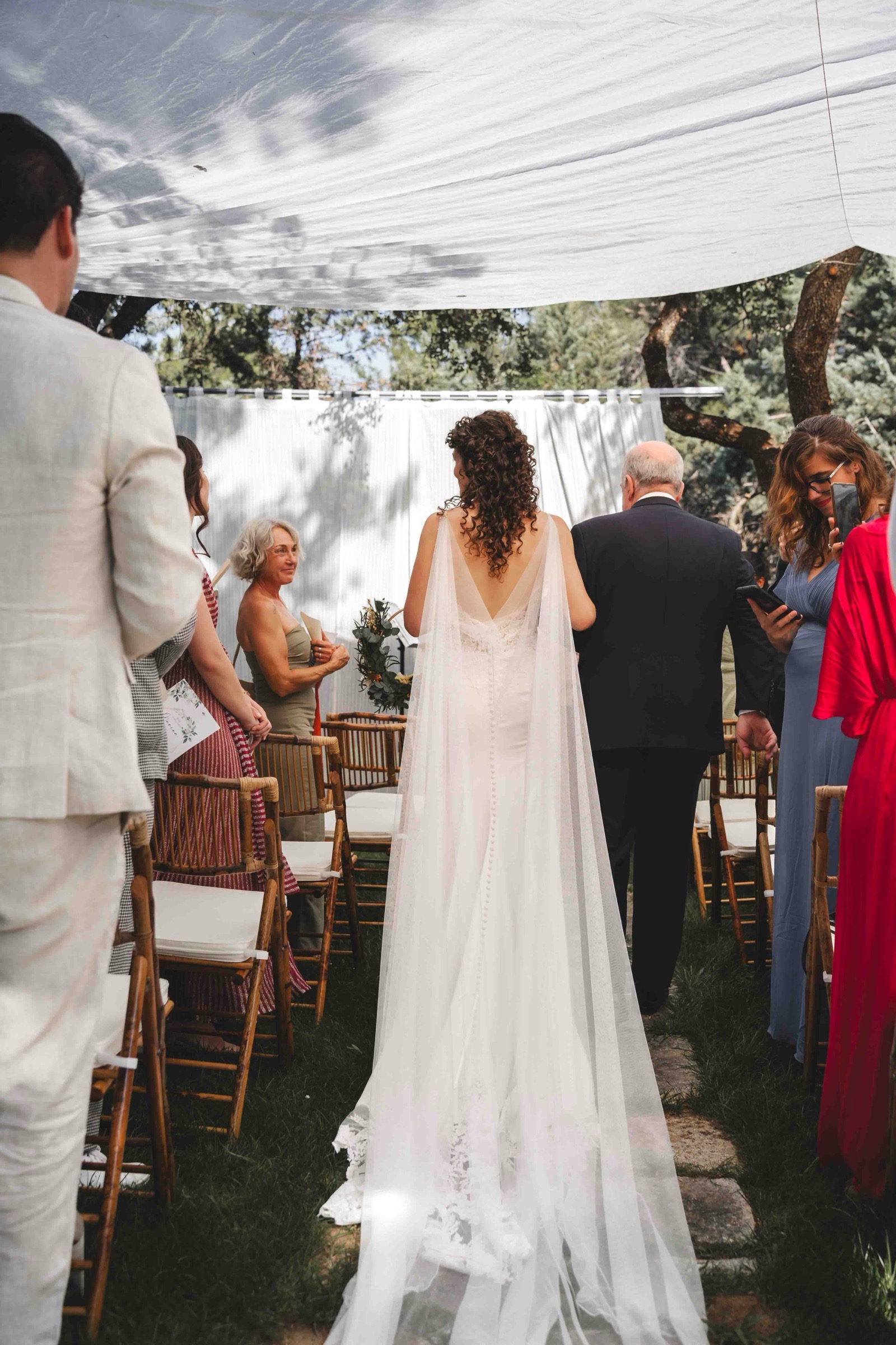 Maria walking down the aisle escorted through seated guests under a white draped canopy surrounded by trees