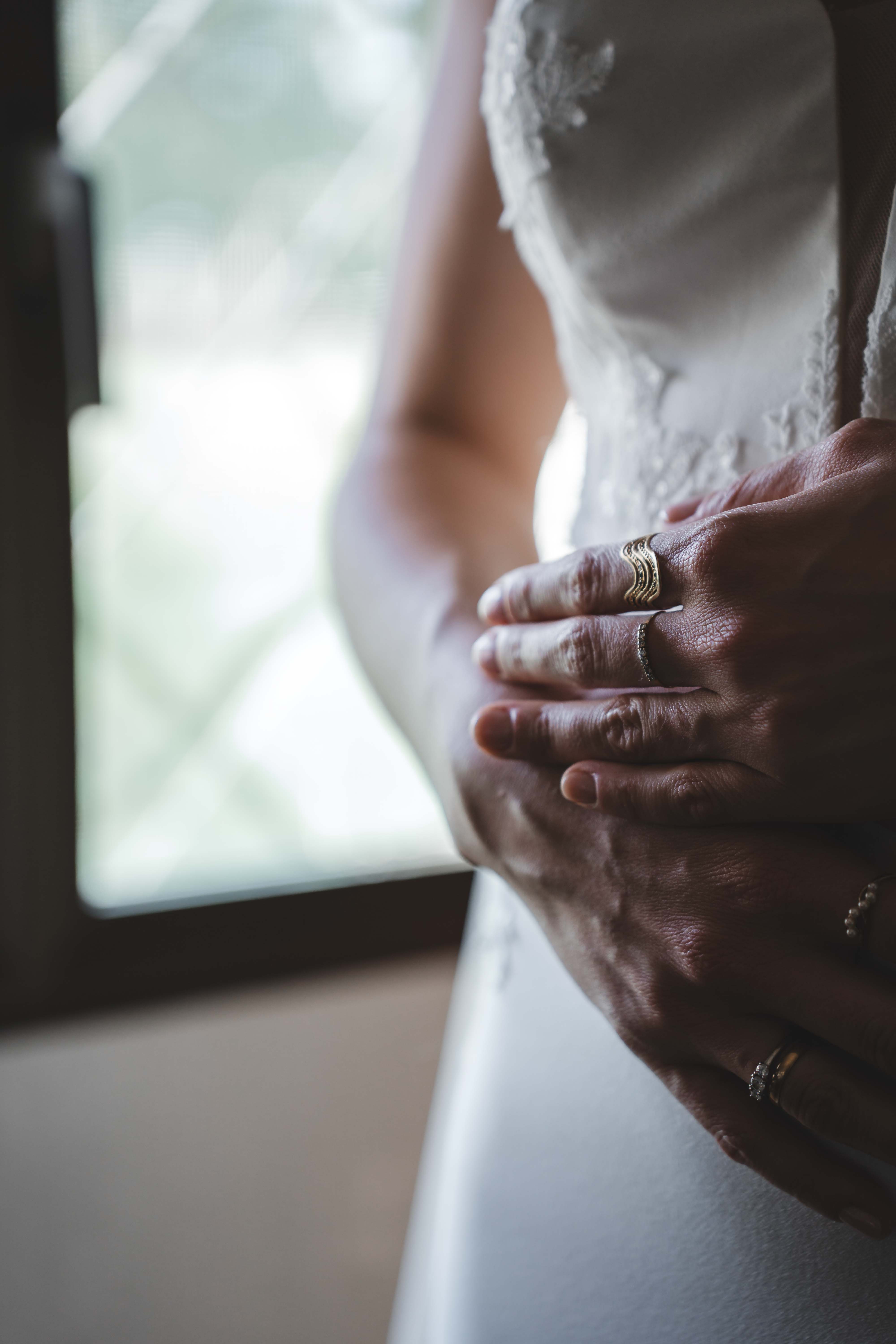 Close-up of hands resting on the wedding dress, rings catching the light by a sunlit window