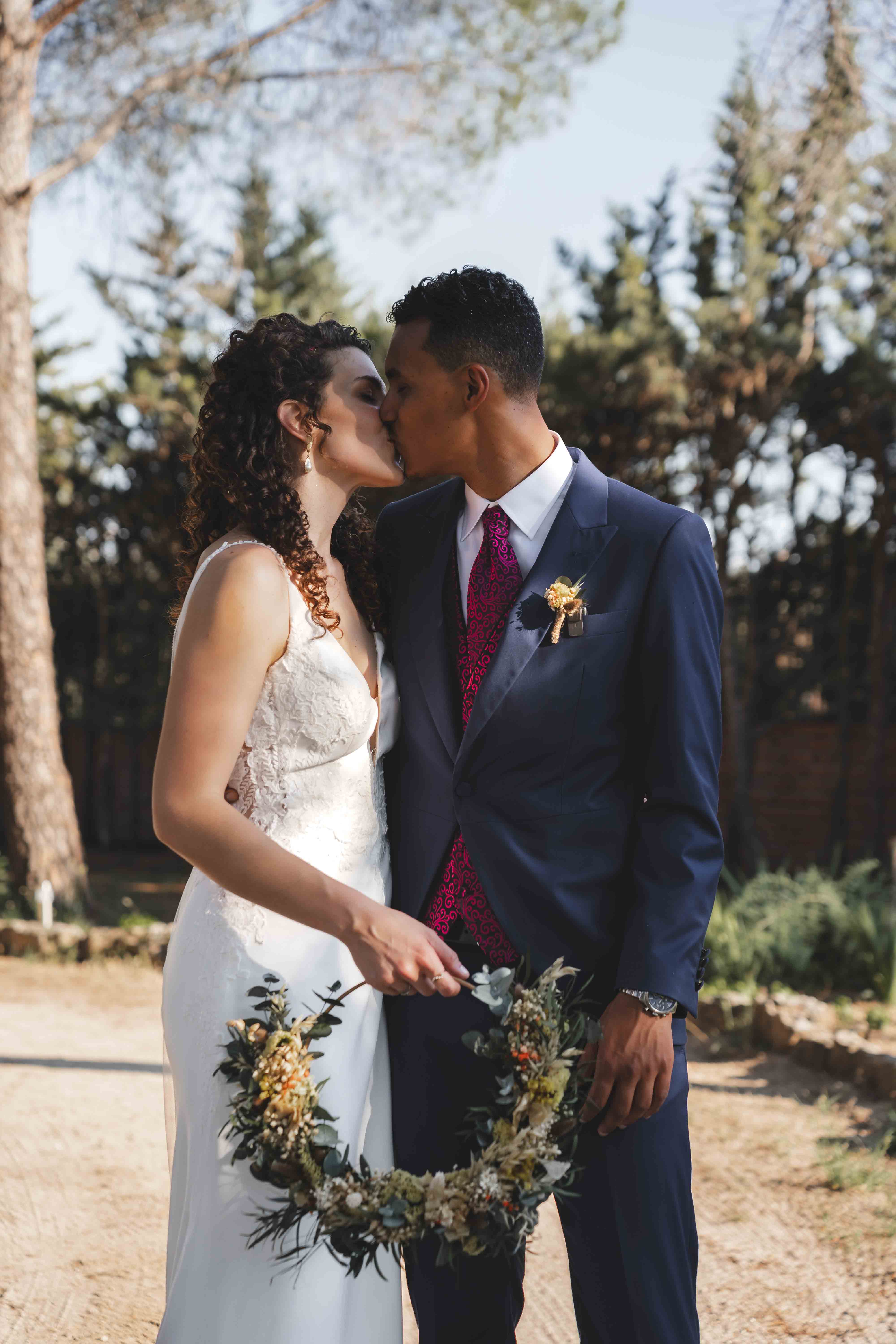 Maria and Leo sharing a kiss, holding the wreath bouquet between them, pine trees in the background