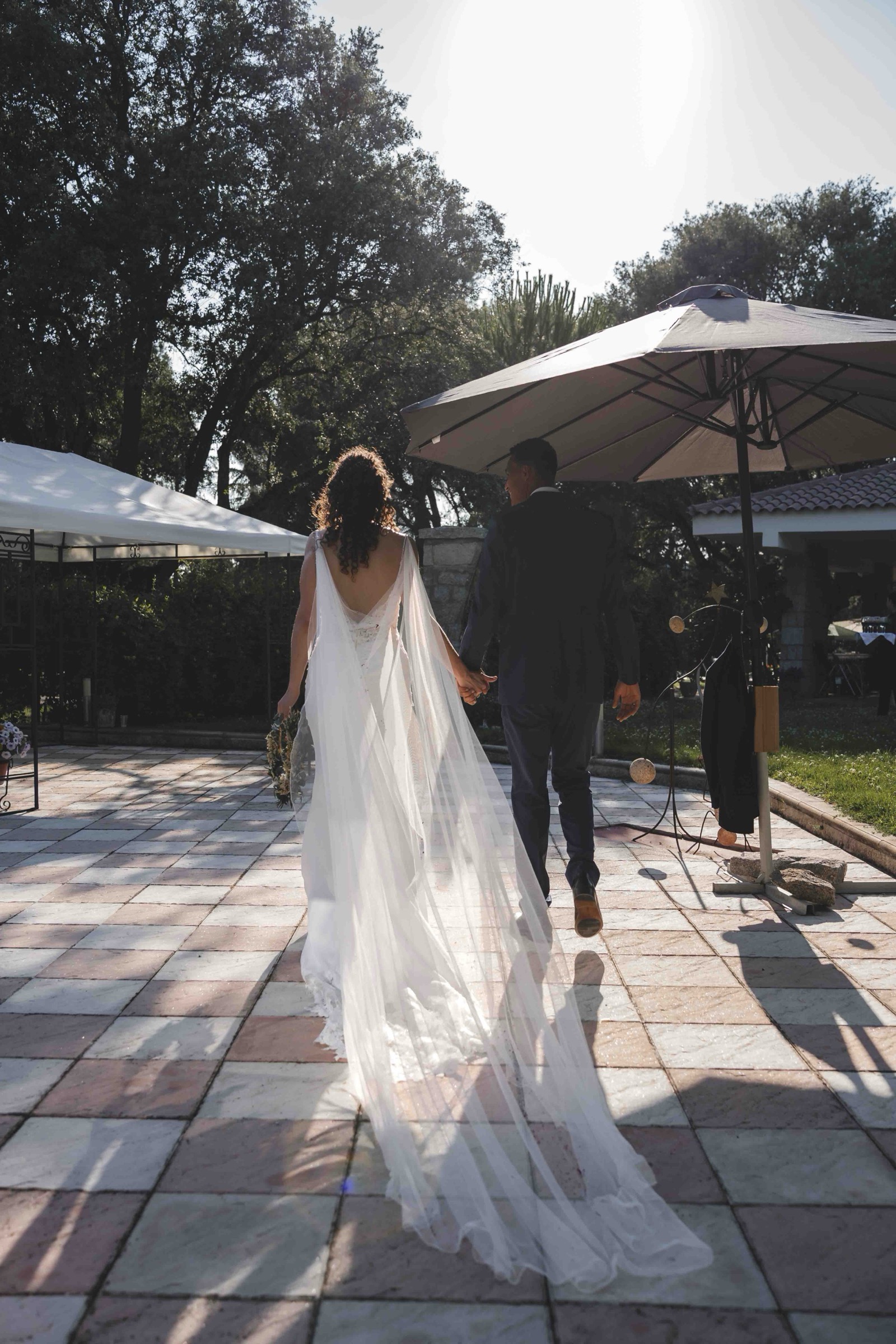 Maria and Leo walking away hand in hand across the patio, backlit by the afternoon sun, veil trailing behind